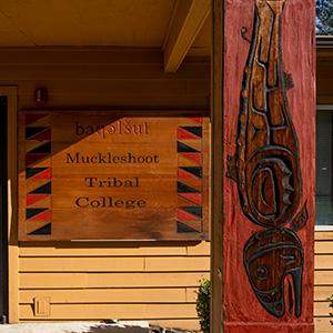 Picture of wood Muckleshoot Tribal College sign. The wood is dark brown. The sign is half in sunlight and half in shadow. In the foreground is a wood carving of a fish.