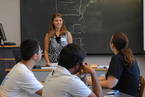 Teacher standing in front of chalkboard smiling at three students seated in front of them