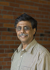 Rajendra Katti wearing a plaid shirt and standing in front of a brick wall