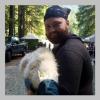 a photo of Tyler Hall. They have a beard and are wearing a blue bandana on their head and a black hoodie. They are holding a white furry animal. 