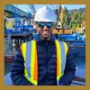 a photo of Vinial Kumar. He is wearing sunglasses, a white hard hat, and a reflective vest over a black coat. He is standing in front of a dock with construction cranes in the background.